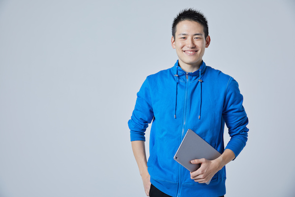 青いジップアップパーカーを着用し、タブレットを手に笑顔で立つ若い男性。シンプルなグレー背景のポートレート