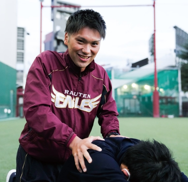 フィットネスジムの店内で、タブレットを持ち笑顔で立つ男性インストラクター。背景にトレーニングマシンが見える