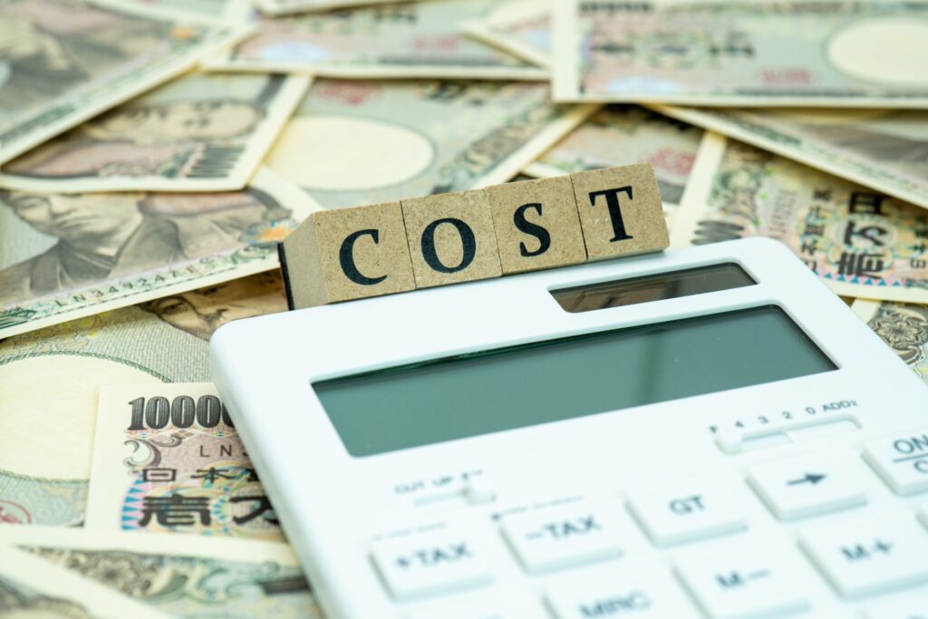 大量の日本円札を背景に、電卓と「COST」の文字ブロックが置かれた写真です。